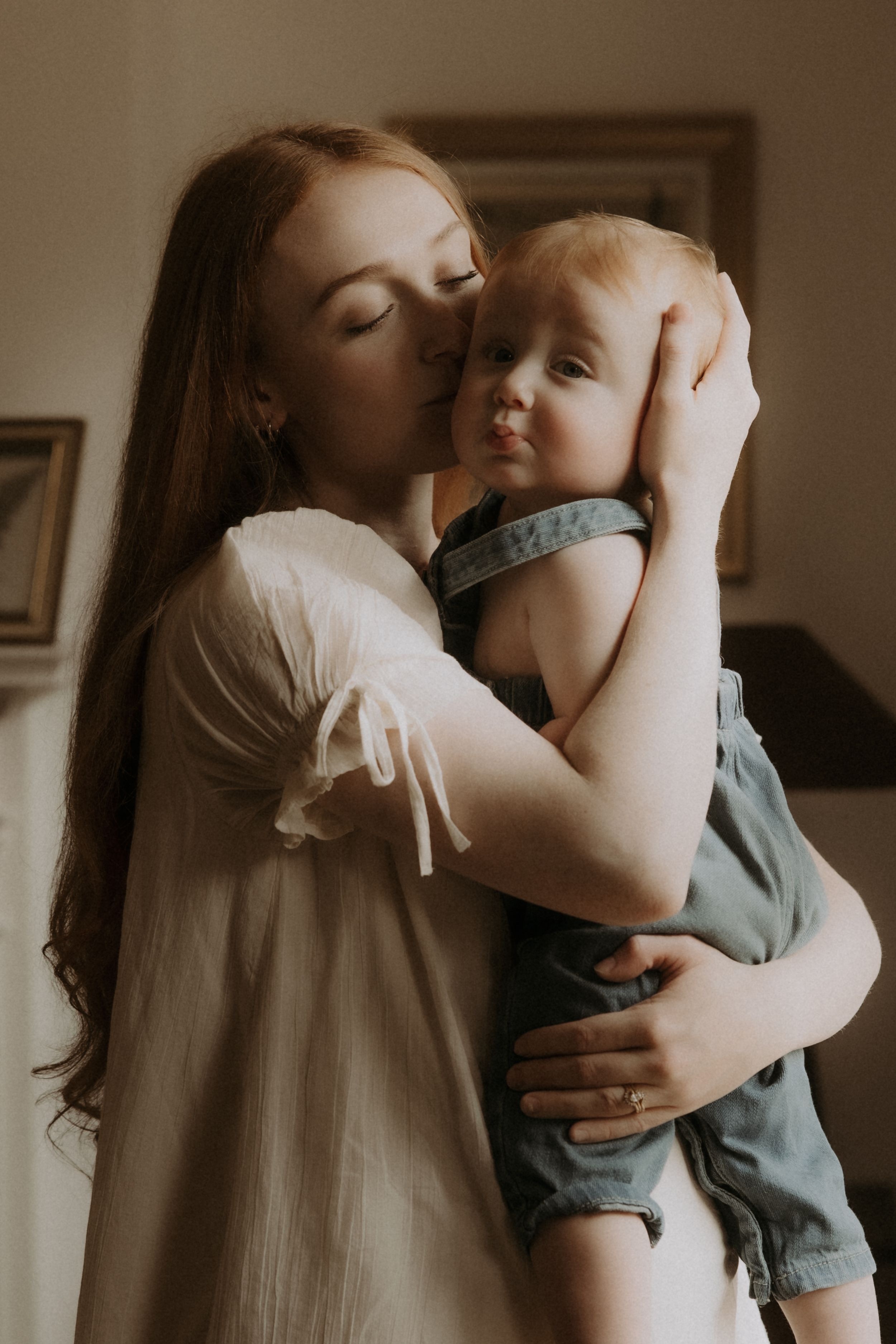 A redheaded mother tenderly kisses her baby boy while holding him close in a warm, softly lit room.