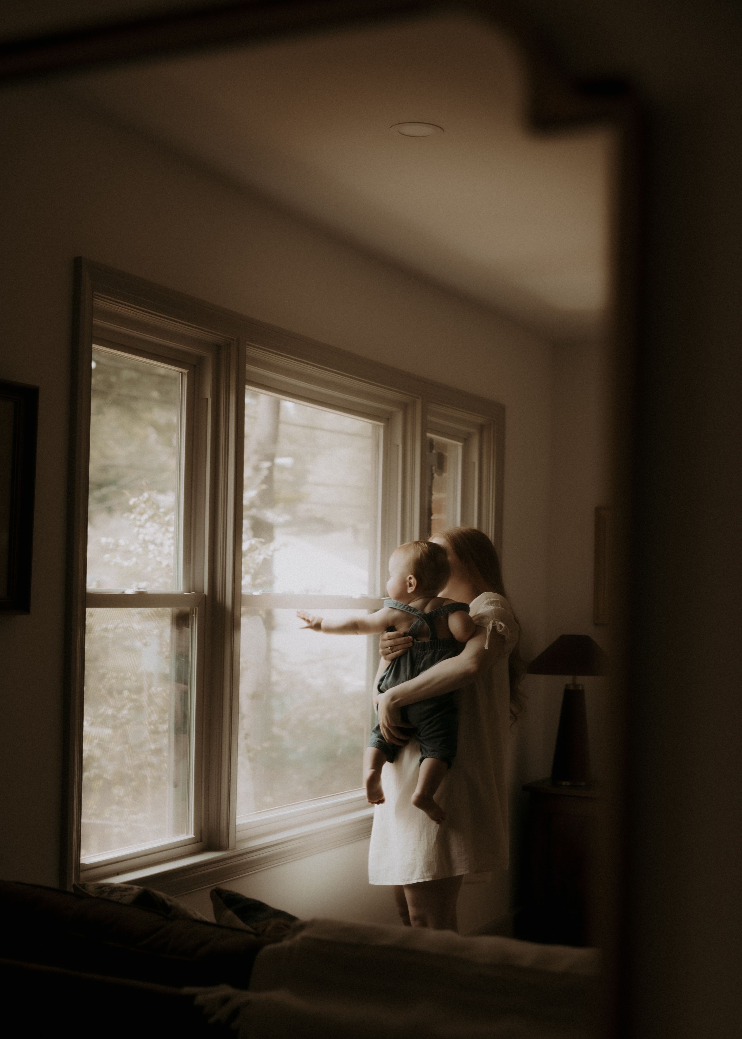 A mother holds her toddler near sunlit windows in a dimly lit room, the child pointing outside with curiosity.