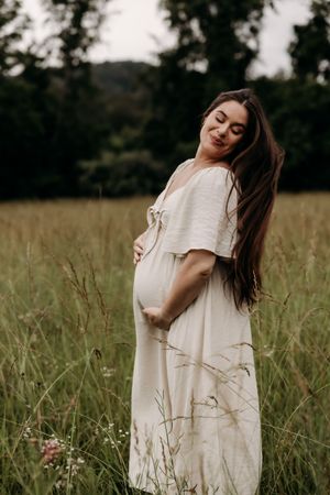 A person in a flowing white dress stands in a sunny meadow of tall grass cradling their pregnant belly.