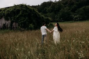 Two people stand together in a golden summer field, their silhouettes framed by tall grass and trees at dusk.