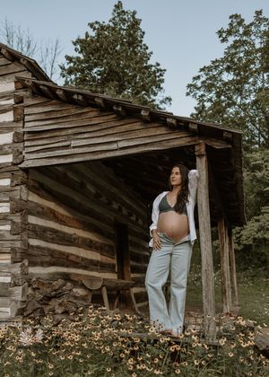 Pregnant woman in white shirt and jeans posing by a rustic log cabin surrounded by wildflowers and trees.