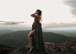 Woman in flowing green dress standing on mountain peak at sunset with scenic valley views.