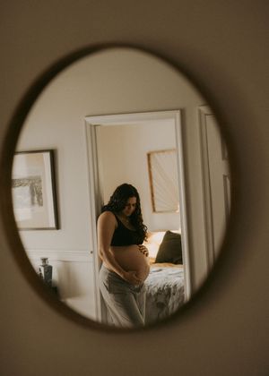 Pregnant woman cradles her baby bump, reflected in a round mirror in a warmly lit home.