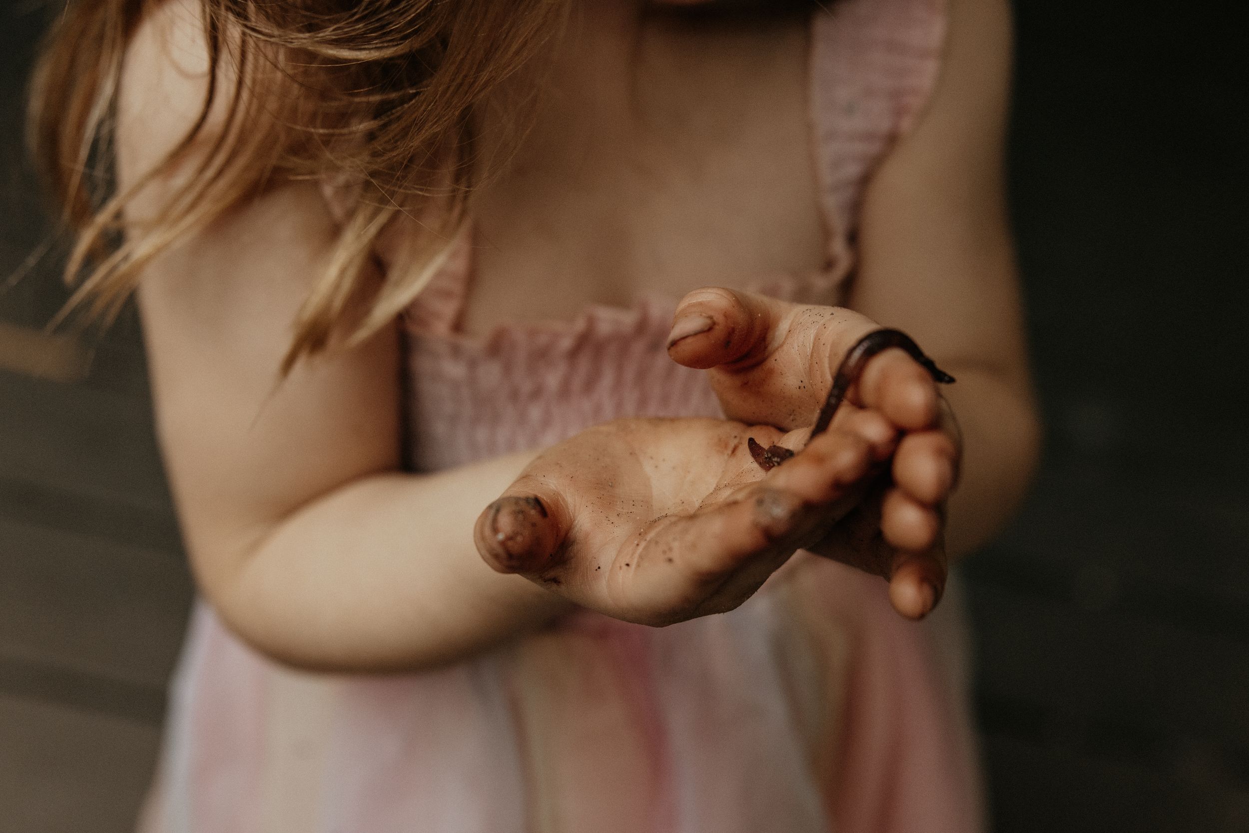 Young girl in pink dress holding out dirty hands, palms up, showing mud-covered fingers outdoors.