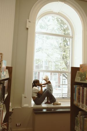 Two people sit together on a window seat in a library, sharing a tender moment among bookshelves.