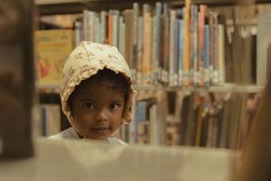 Young child wearing a floral bonnet browsing books in a library, peeking curiously between colorful bookshelves.