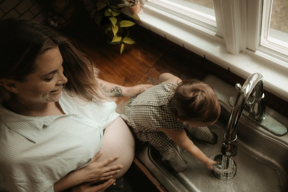 Pregnant woman smiling while toddler plays at kitchen sink near window in cozy home setting.
