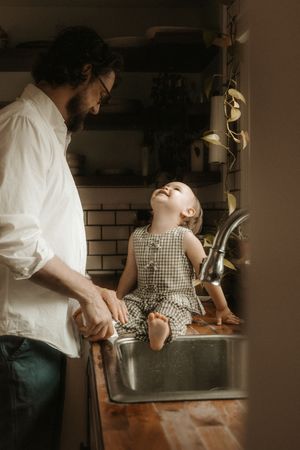 A bearded father watches his toddler daughter sitting in a kitchen sink, surrounded by plants in a cozy home.