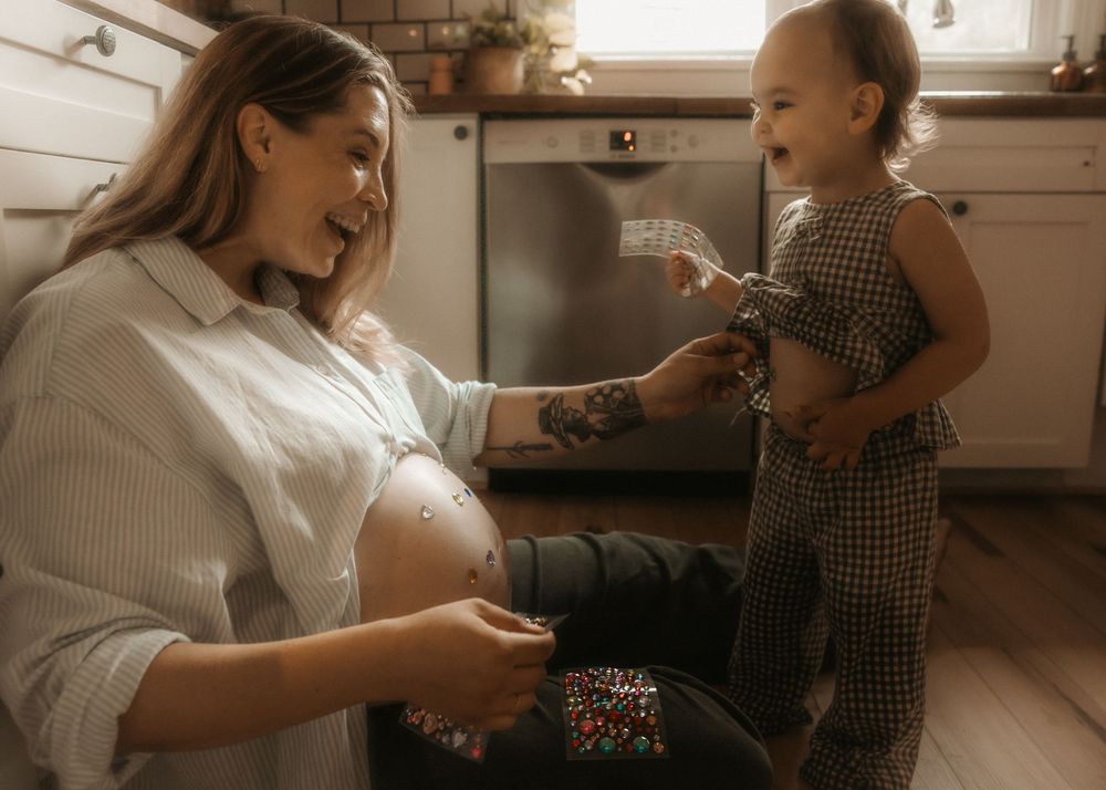 Pregnant woman kneeling in kitchen, smiling at toddler holding confetti during gender reveal celebration.