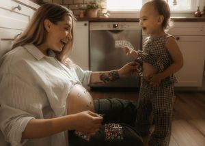 Pregnant woman kneeling in kitchen, smiling at toddler holding confetti during gender reveal celebration.