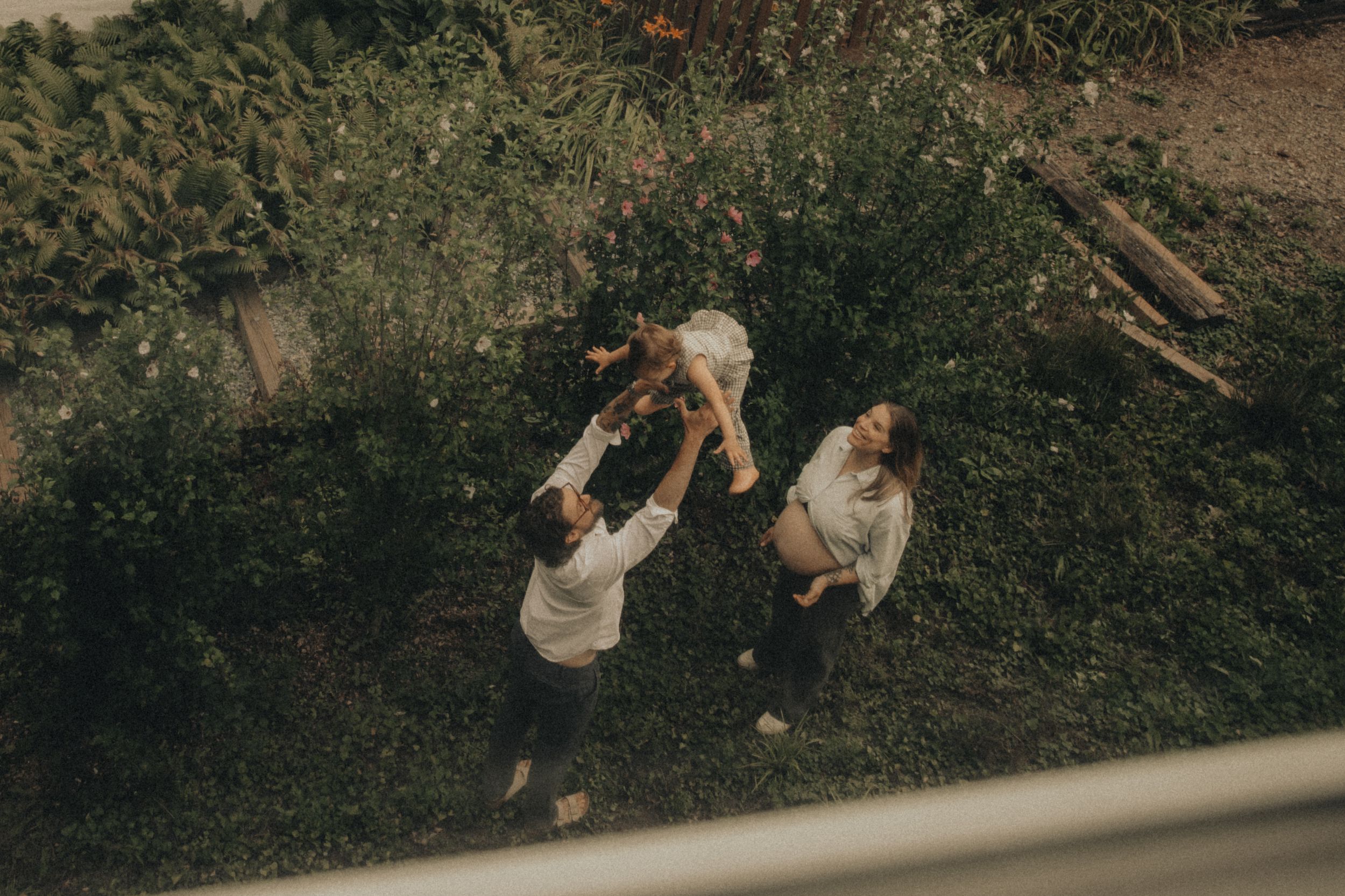 Aerial view of two adults lifting a child in a lush green garden outdoors.