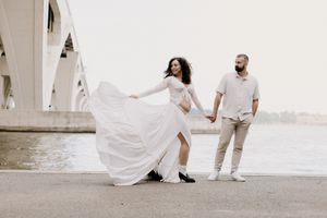 A romantic couple in wedding attire poses along a waterfront as the bride's white dress flows gracefully in the breeze.