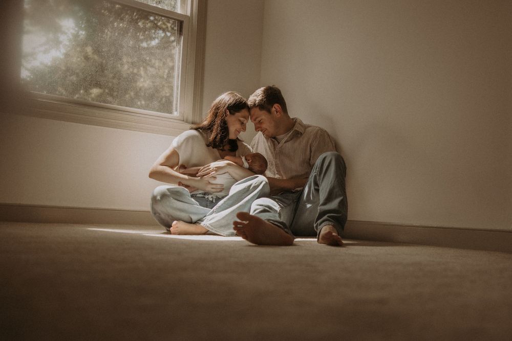 Couple sitting on floor by window, sharing tender moment with newborn baby in soft natural light.