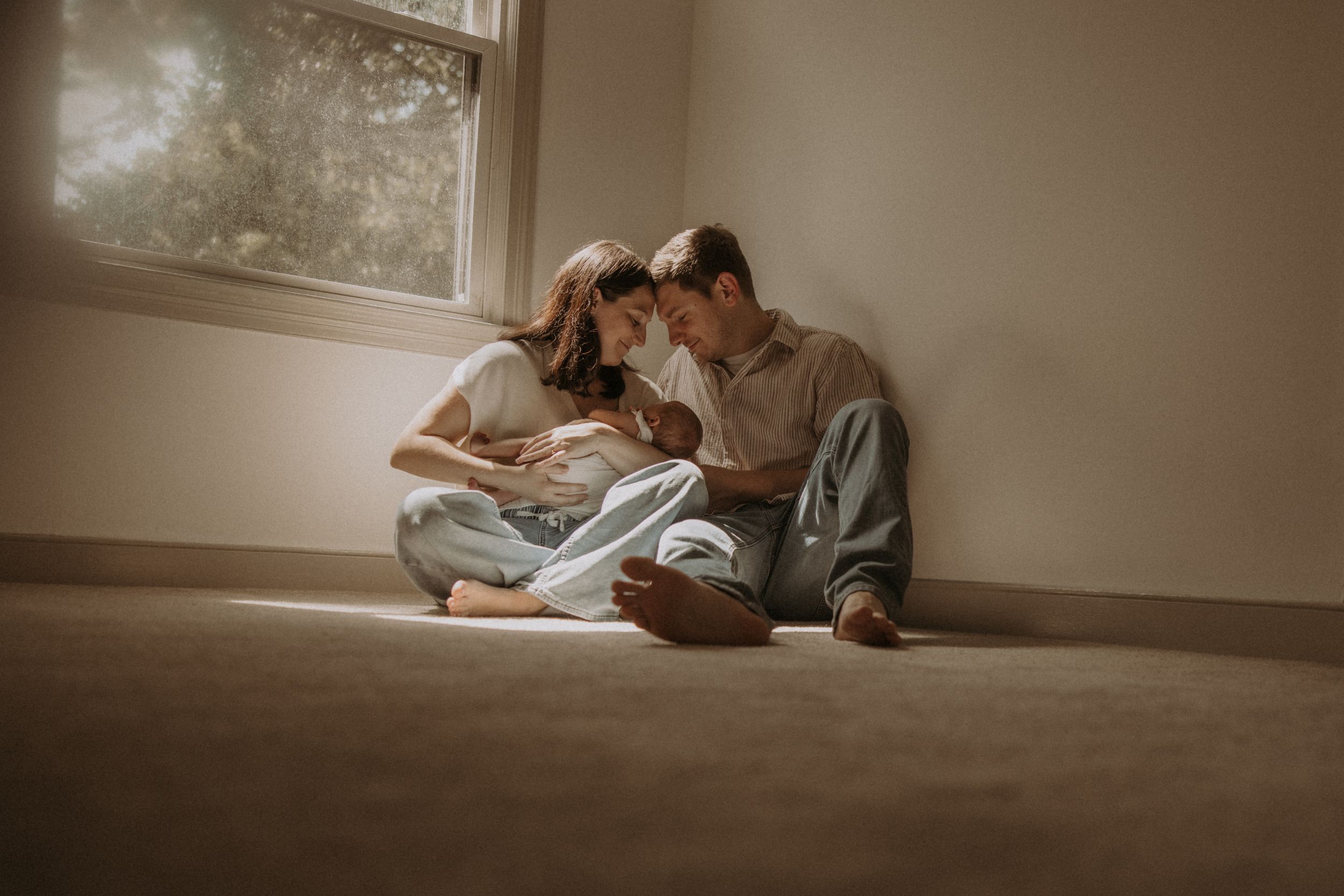 Couple sitting on floor by window, sharing tender moment with newborn baby in soft natural light.