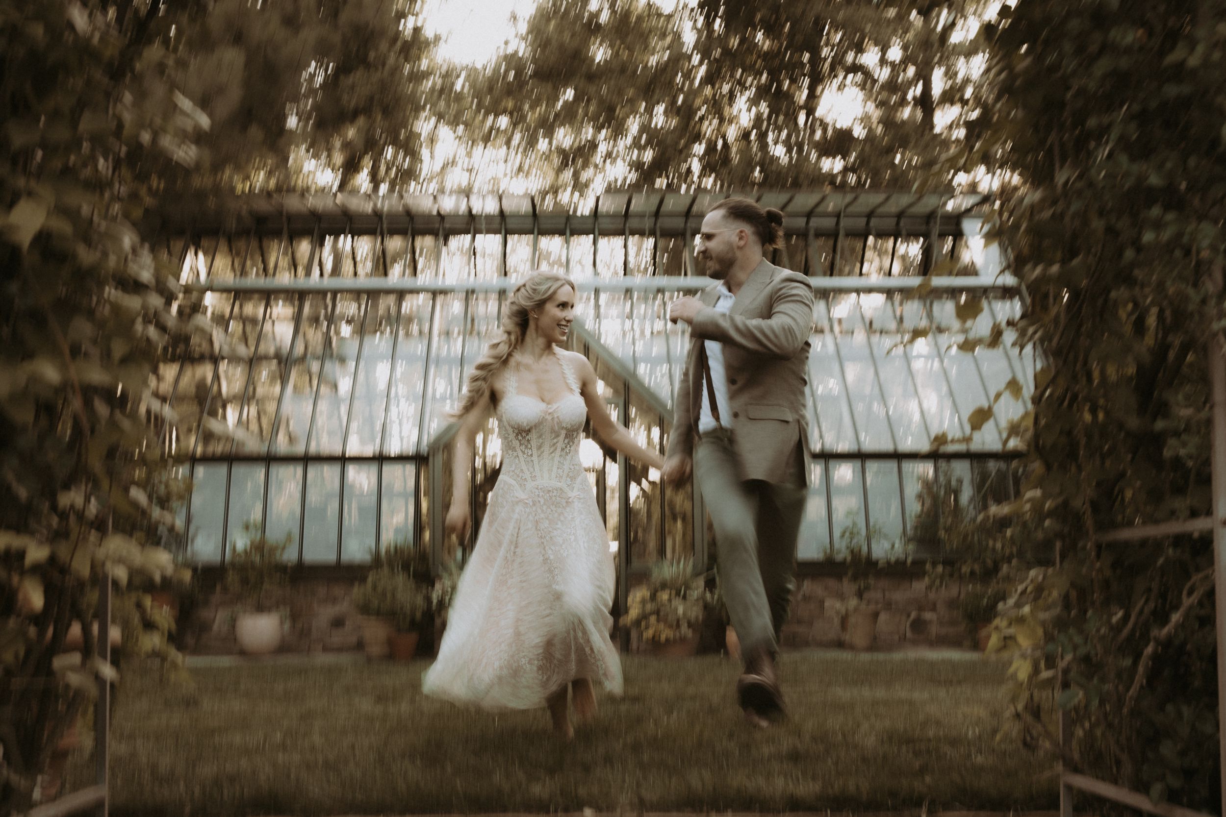 Bride and groom joyfully running hand in hand in a lush garden with a greenhouse backdrop.