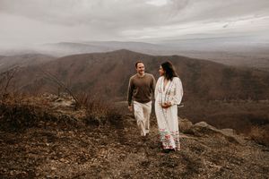A couple in white outfits stands together on a scenic mountain overlook with sweeping valley views in the distance.