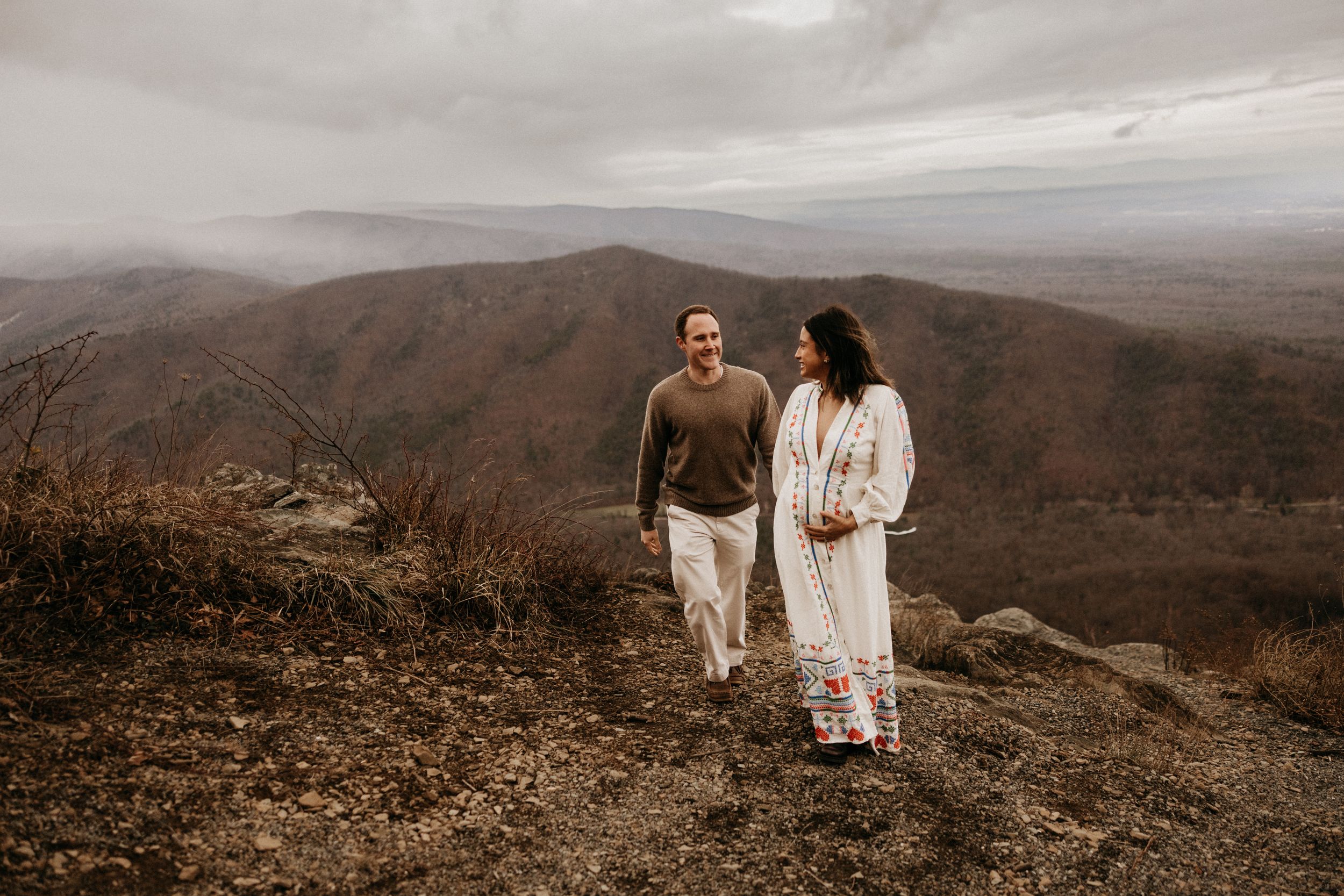 A couple in white outfits stands together on a scenic mountain overlook with sweeping valley views in the distance.