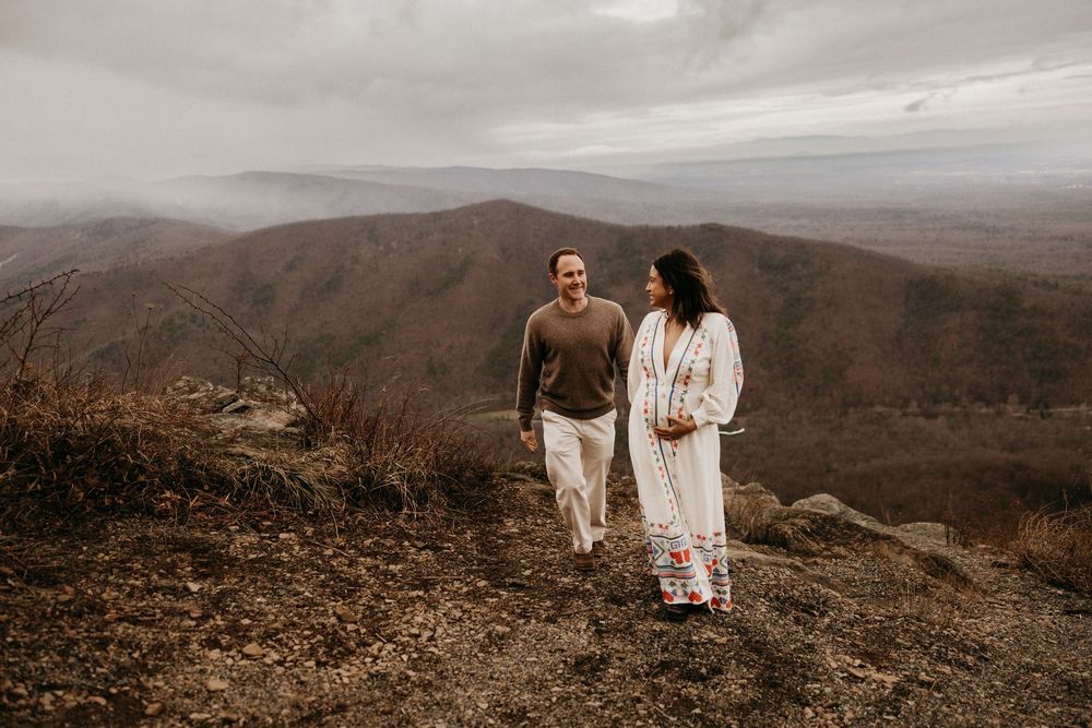 A couple in white outfits stands together on a scenic mountain overlook with sweeping valley views in the distance.