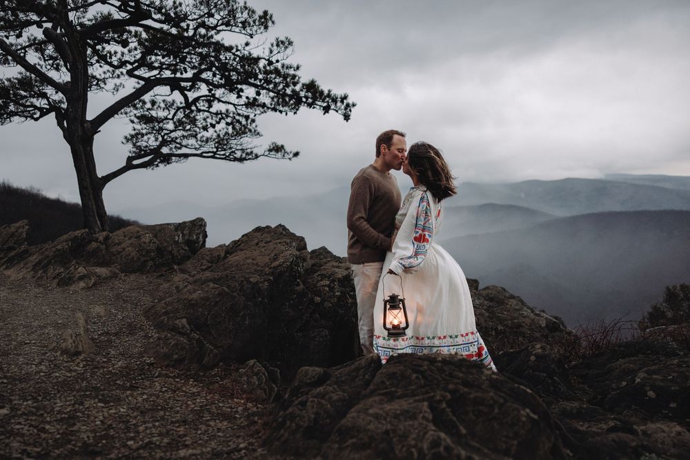 Couple sharing an intimate moment on a rocky mountain overlook at sunset with dramatic mountain views in the background.
