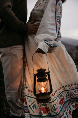 A person wearing an embroidered white dress holds a glowing vintage lantern at dusk, creating a moody atmospheric scene.
