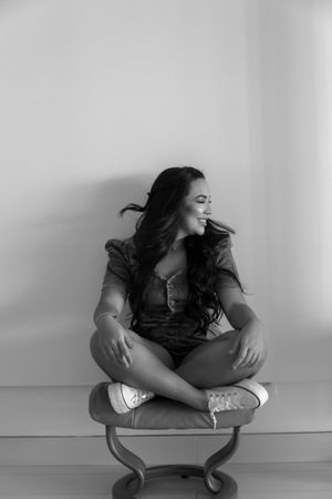 A candid portrait in black and white shows someone sitting cross-legged on a small curved chair against a plain wall.