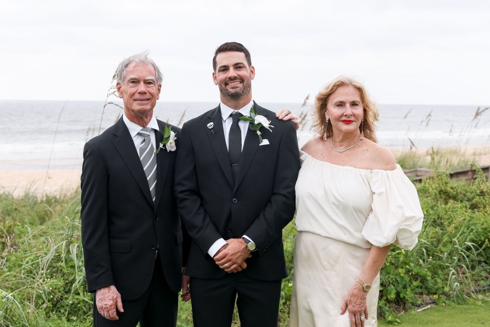Three elegantly dressed people pose together by the ocean on a formal occasion with coastal scenery in the background.