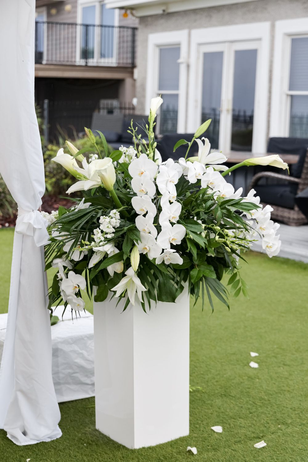 Large white floral arrangement with lilies and greenery displayed on a modern white pedestal outdoors.