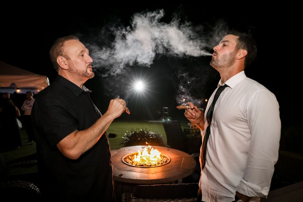 Two men enjoy cigars while standing near an outdoor fire pit at night.