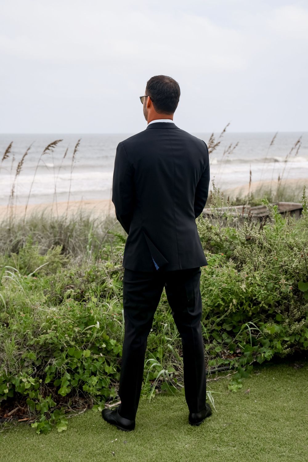 A groom in a black suit looks out over the ocean from a coastal wedding venue.