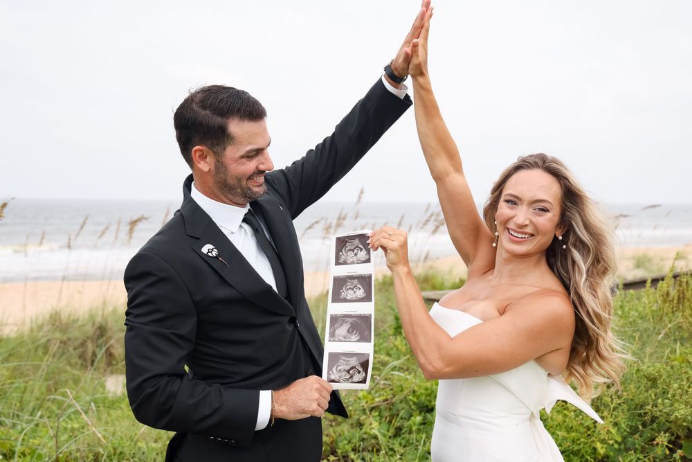 A joyful couple gives each other a high five while holding ultrasound photos by the beach.