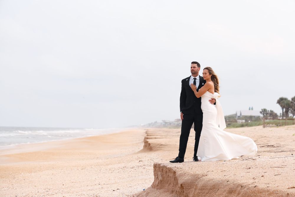 A wedding couple stands on a sandy beach cliff overlooking the ocean.