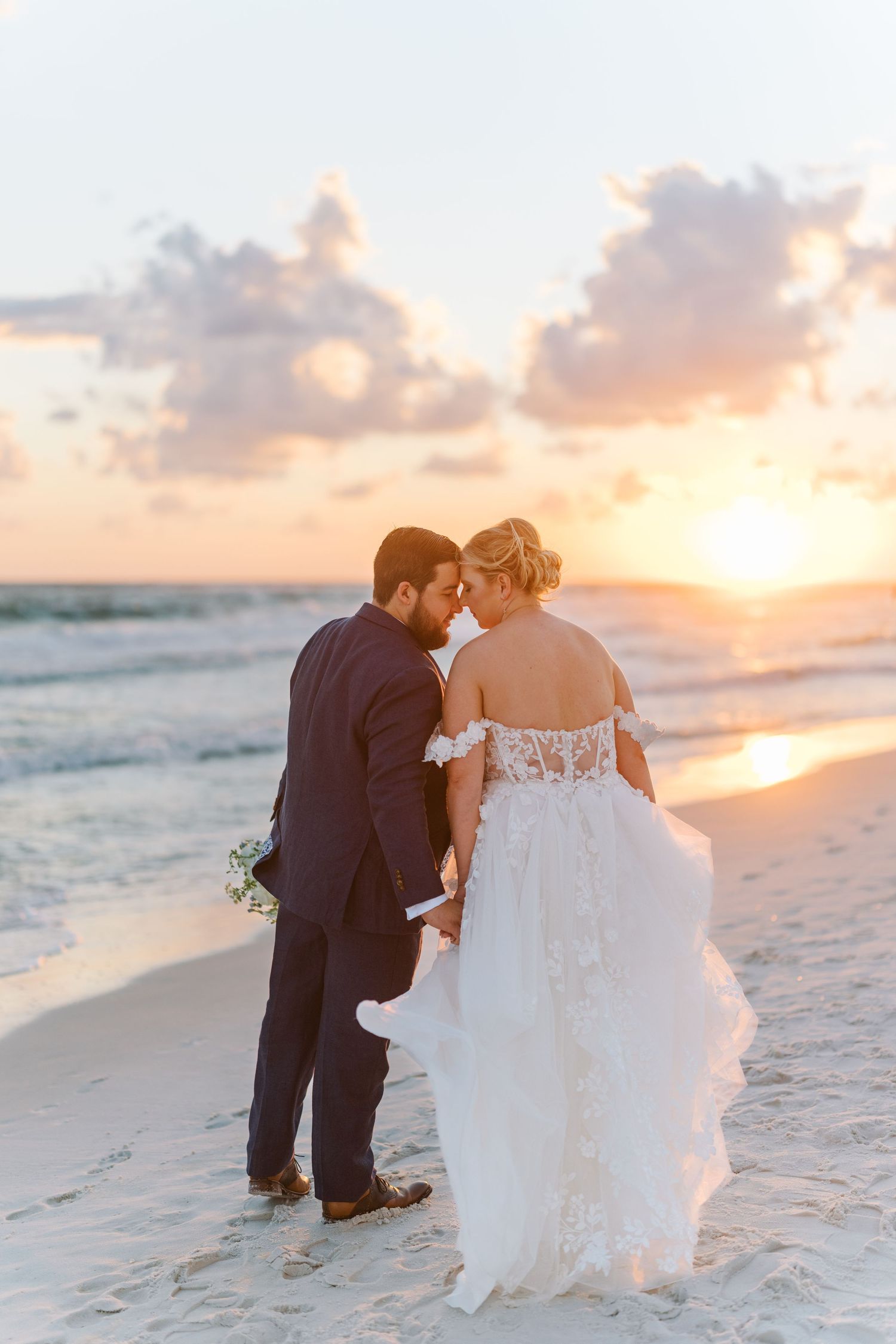 A couple walks along a sandy beach during sunset, her white dress flowing in the ocean breeze.