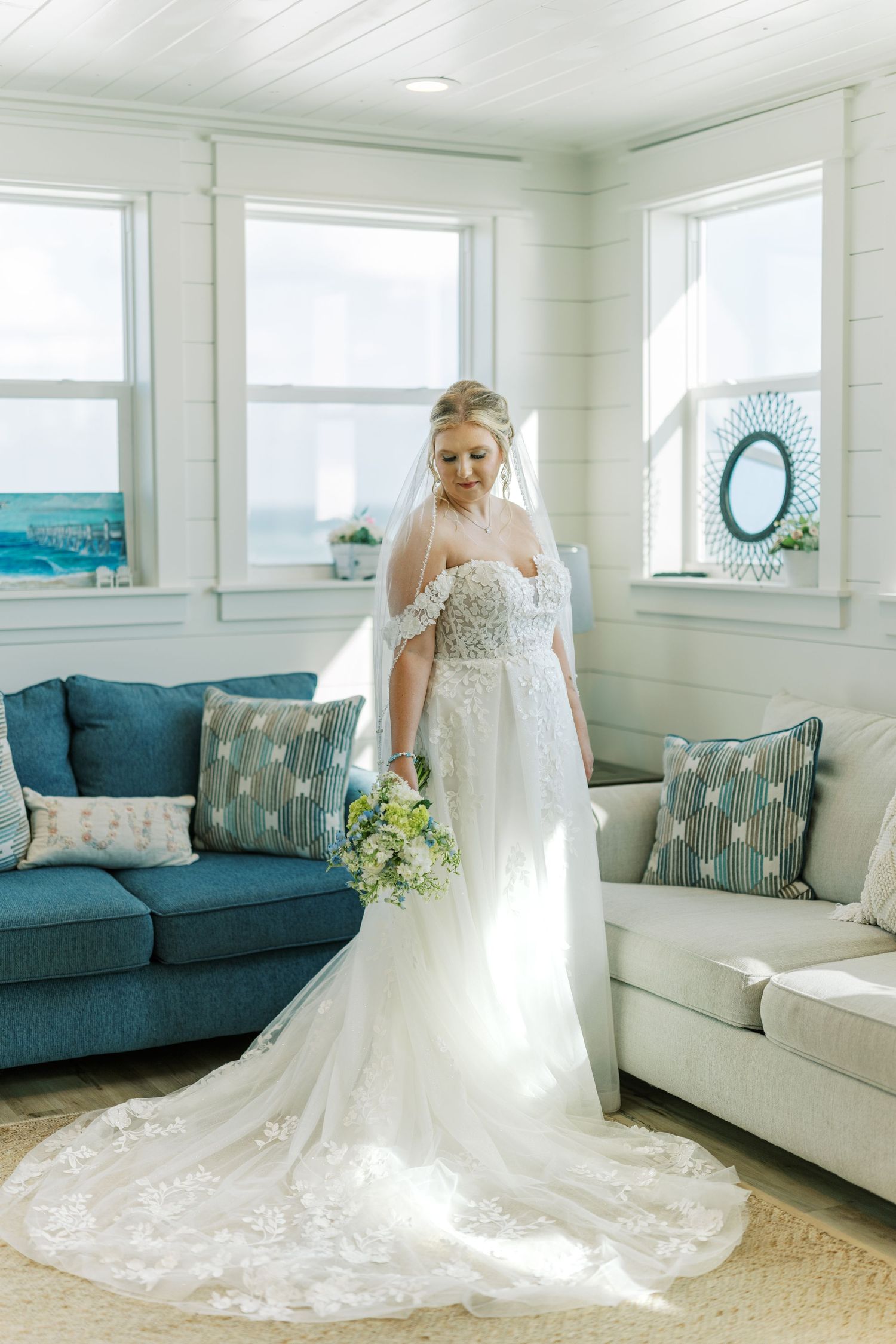A bride in a white lace gown with train poses in a coastal-themed room with blue furniture and ocean views.