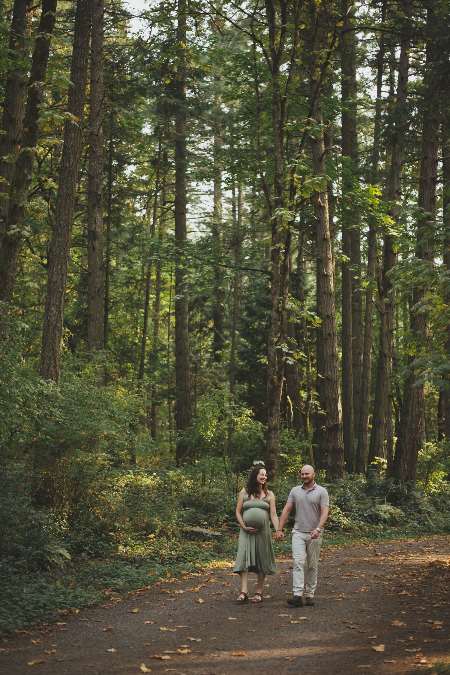 Expecting couple holding hands and walking along a forest trail surrounded by tall trees.