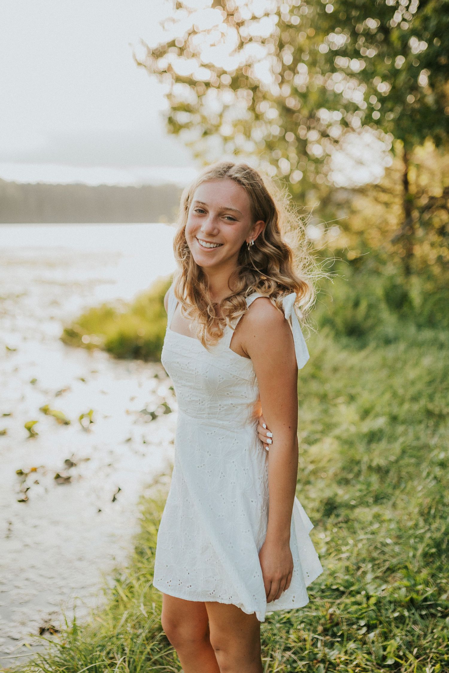 A child in a white summer dress plays outdoors at sunset in a grassy field near the water.