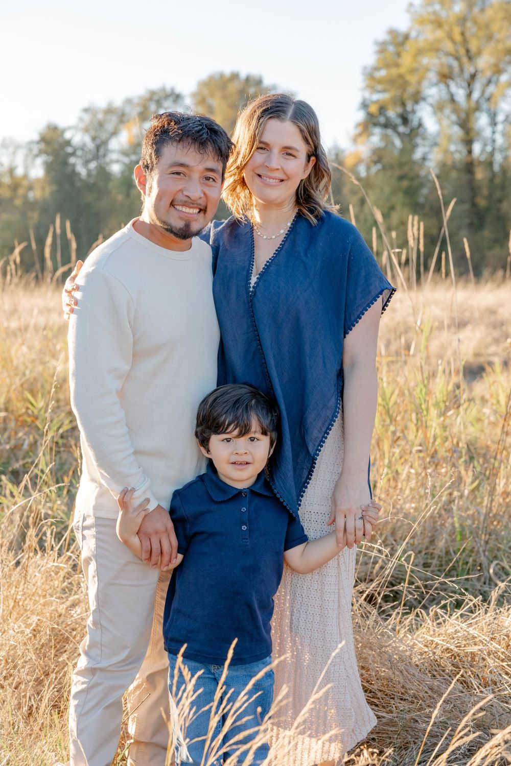 Happy family of three posing in golden field, parents smiling with young son in matching blue outfits.