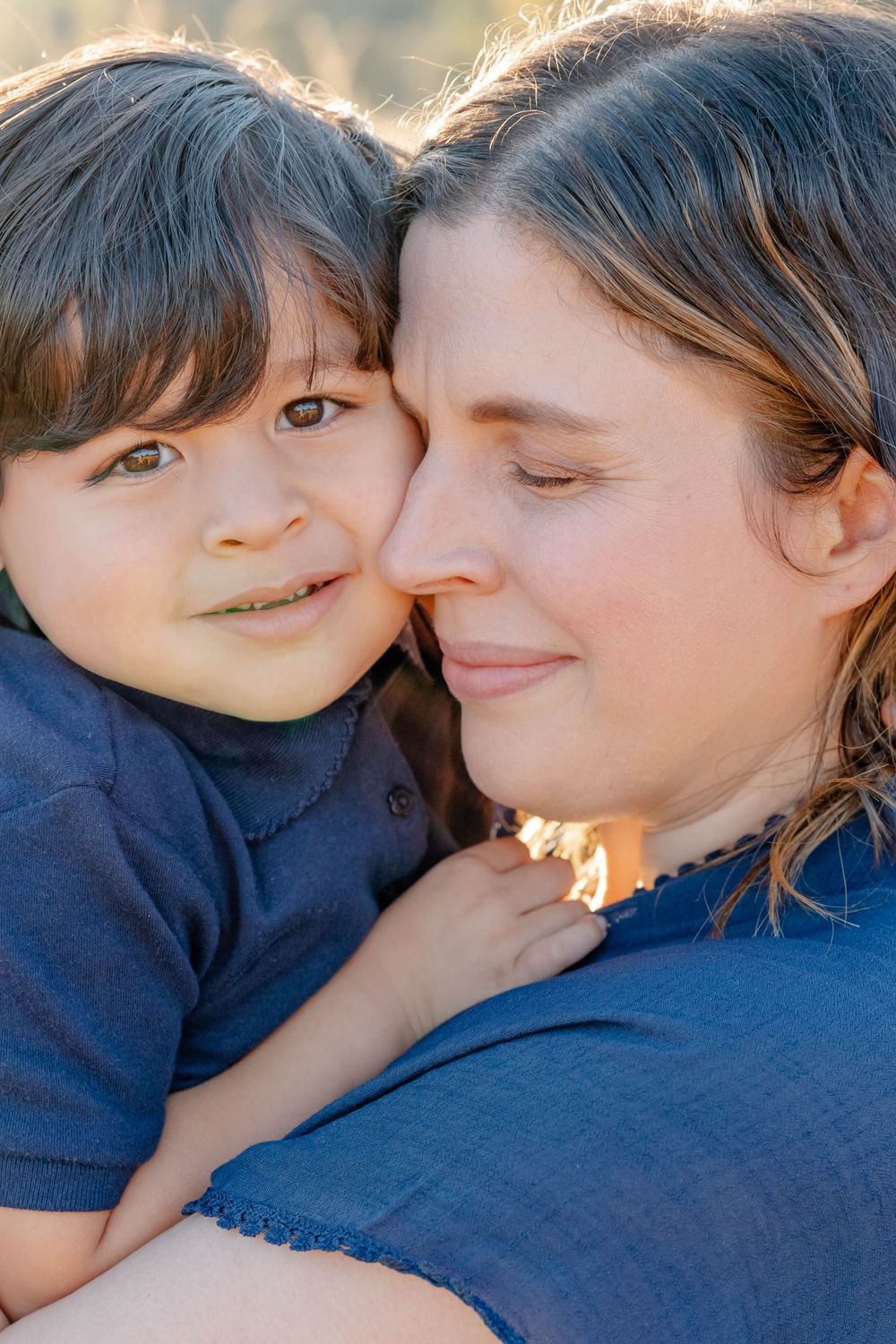 Close-up of loving mother holding young son cheek-to-cheek, both smiling warmly in soft sunlight.