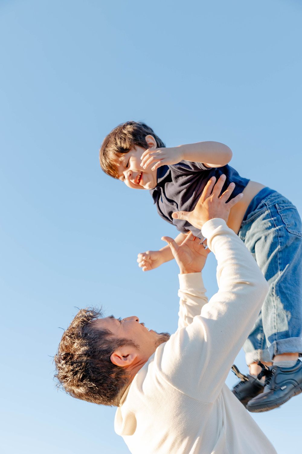Father joyfully lifting young son high against clear blue sky, both laughing during outdoor playtime.