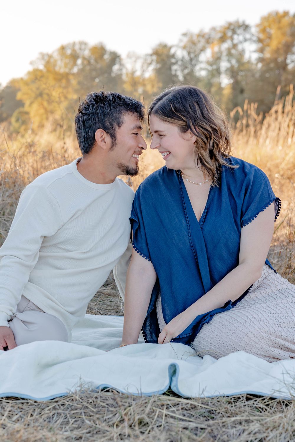 Couple sitting close on white blanket in golden field, smiling at each other in warm sunset light.