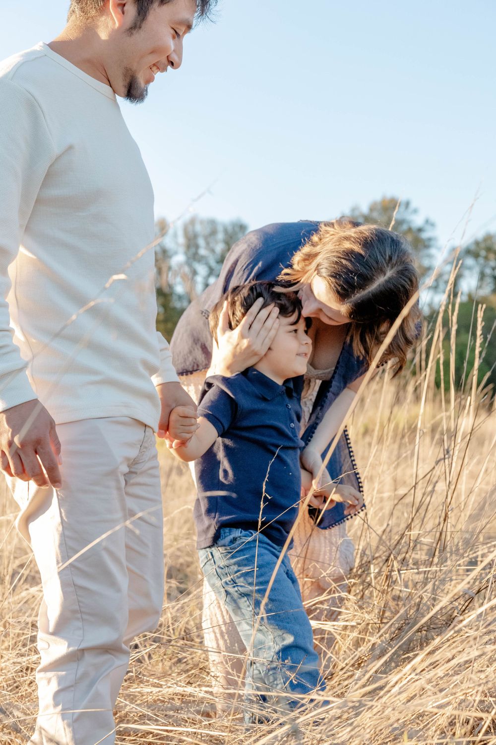 Mother tenderly brushing sons hair in sunny golden field while father stands nearby watching warmly.