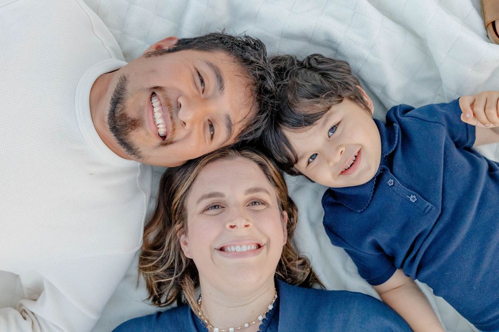 Happy family of three lying on white blanket, smiling up at camera in a top-down portrait shot.