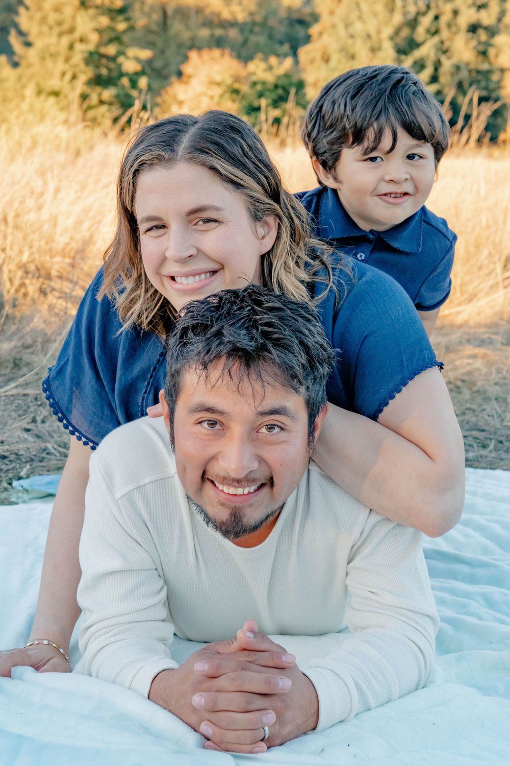 Playful family portrait with son perched on parents backs while lying on blanket in golden field.