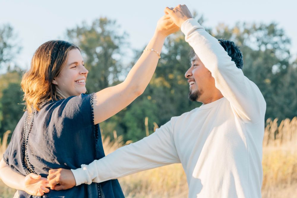 Couple dancing joyfully in a sunny field, holding hands raised, smiling at each other outdoors.