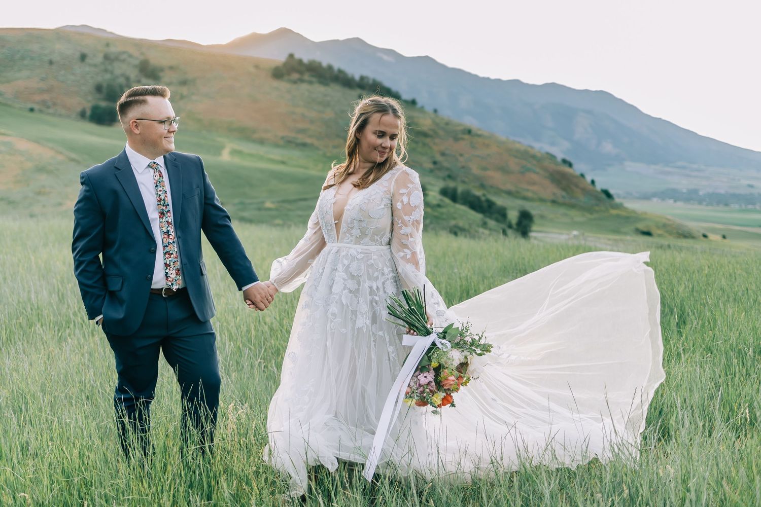 Wedding couple holding hands in mountain meadow with bride's dress flowing in the wind.