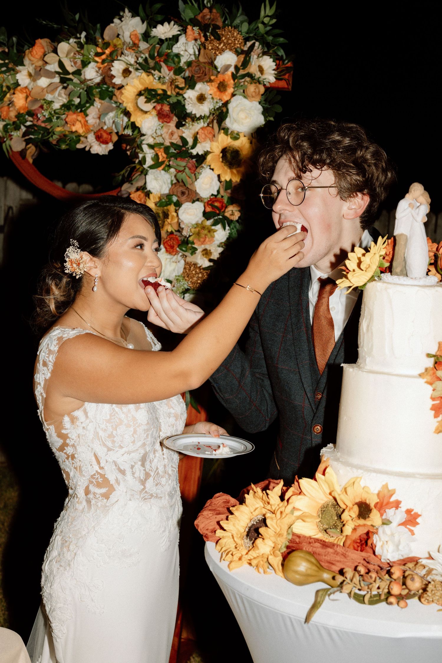 Wedding couple feeding each other cake in front of sunflower decorations and tiered white cake.