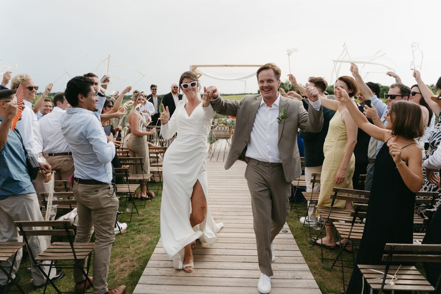 Wedding couple jubilantly walks down a wooden boardwalk aisle while guests cheer and celebrate.