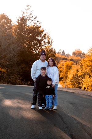 Young family of four stand together between autumn trees on road Cottonmill Park, Kearney, NE.