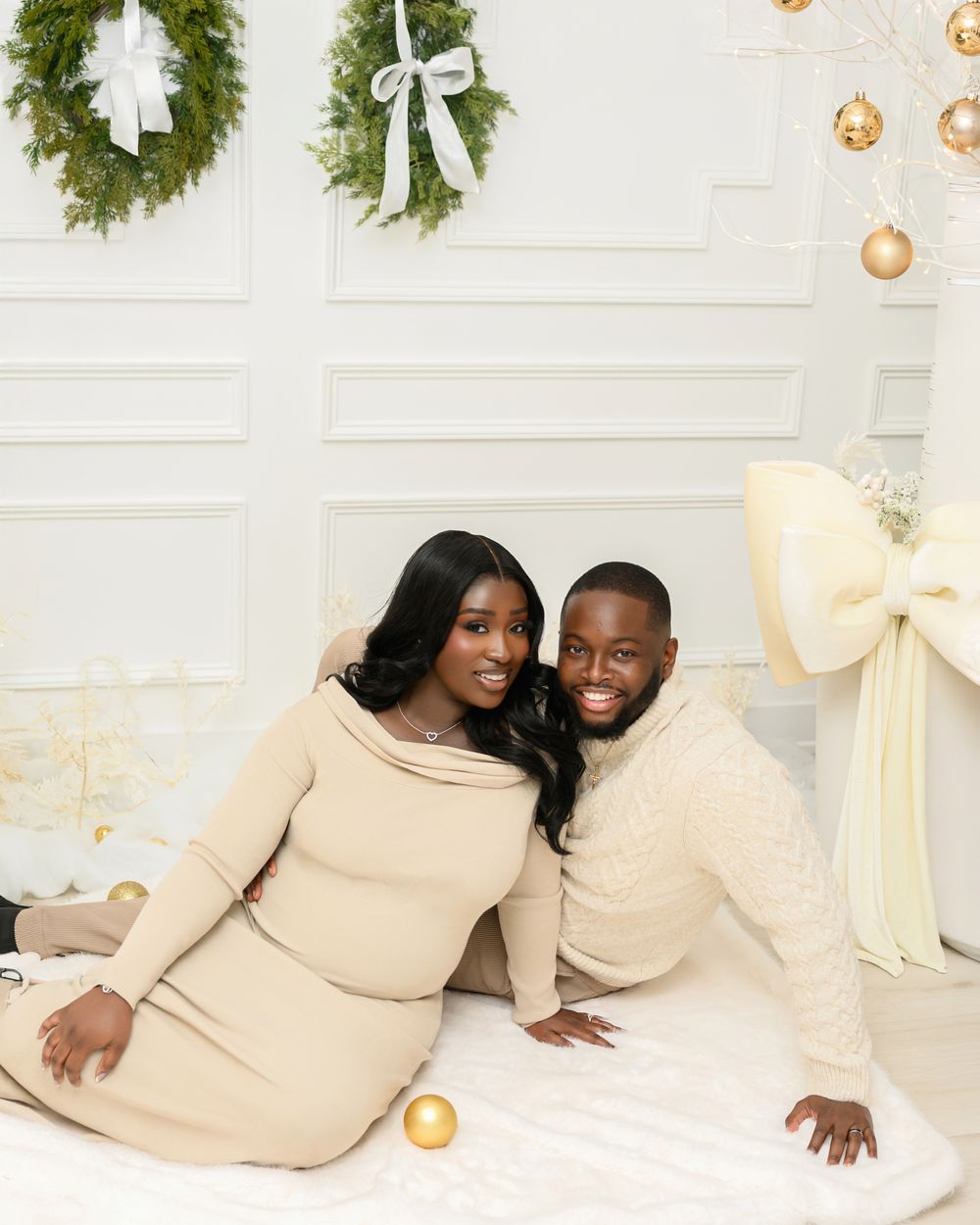 Elegant holiday photo setup with cream colored outfits against white wall decorated with evergreen wreaths and gold ornaments.