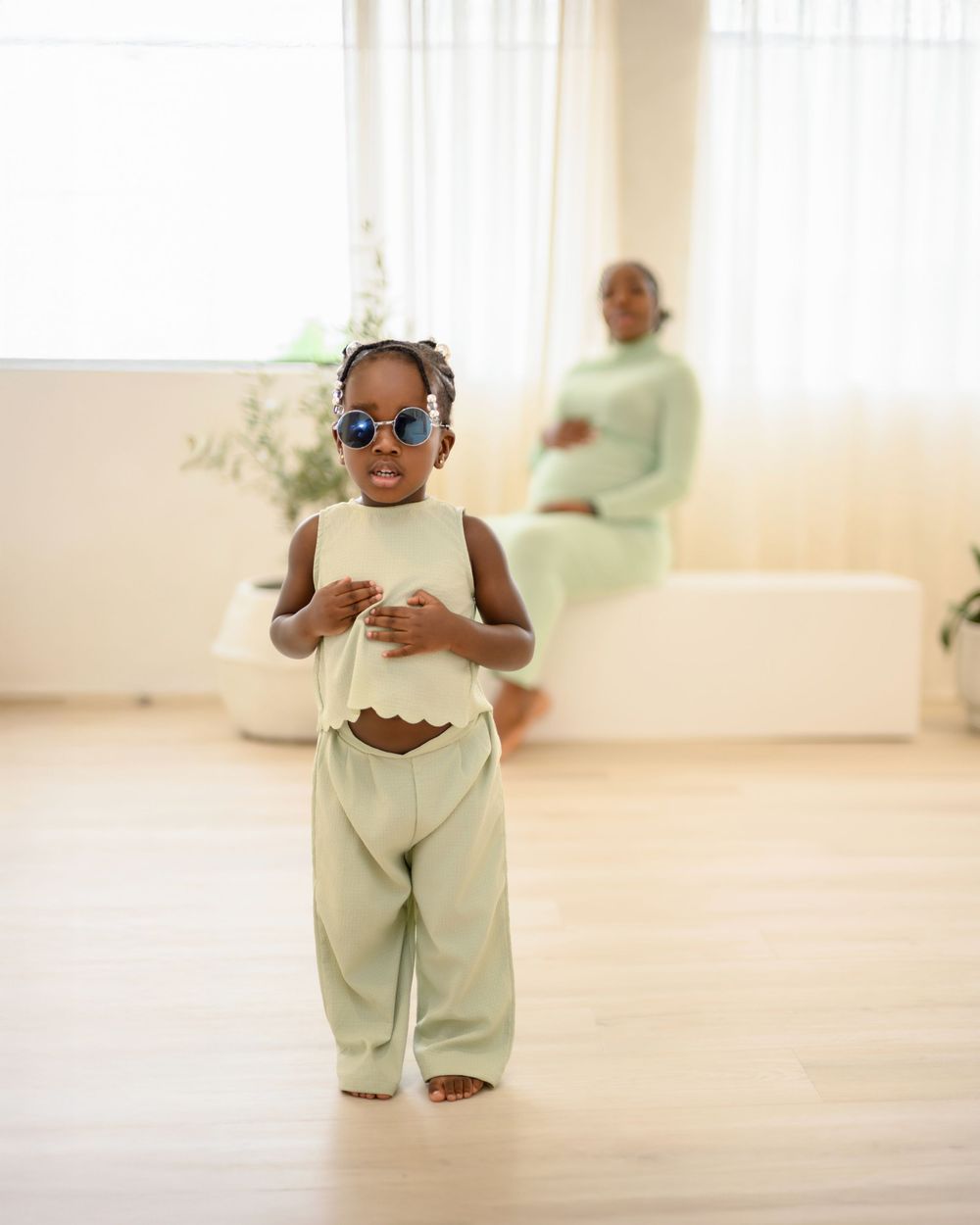 A small baby wearing sunglasses and cream-colored outfit dances in a bright sunlit room.