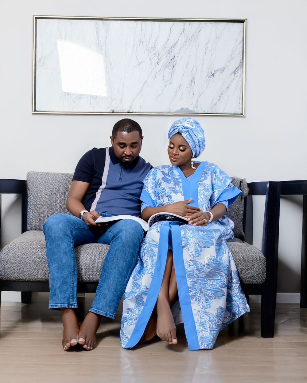 Two people sit comfortably on a gray couch while reading or looking at their phones against a light marble wall.